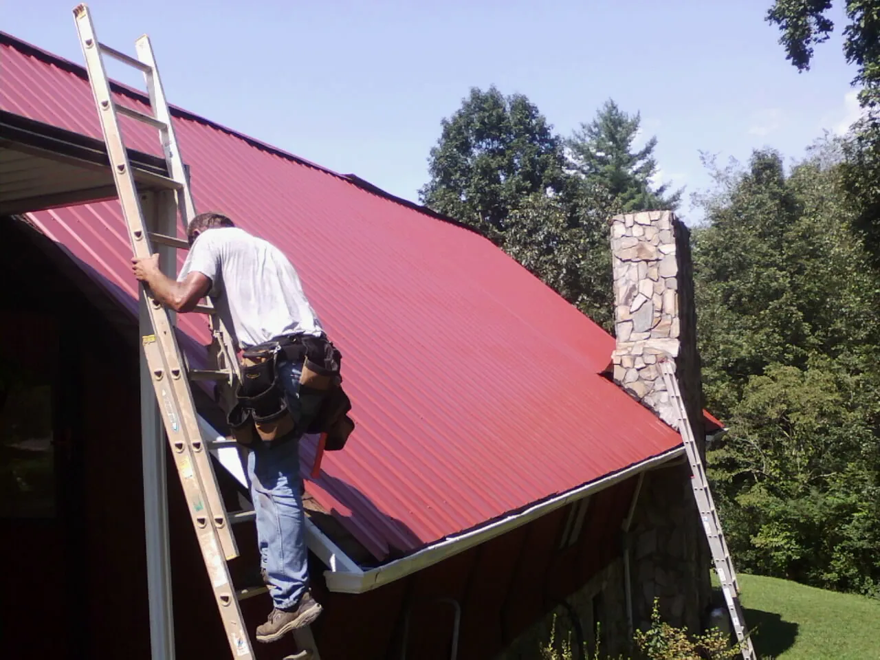 Roofer on a ladder beside a red metal roof and stone chimney — Soffit & Fascia Repair by Roofing Brothers in Kellogg
